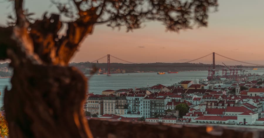 Beautiful sunset over Lisbon with a view of the iconic 25 de Abril Bridge, framed by trees.